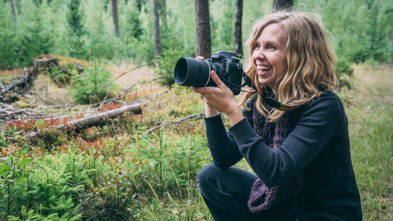 Fotogfafen Maria sitter på huk i skogen med sin kamera beredd för att fotografera och ler