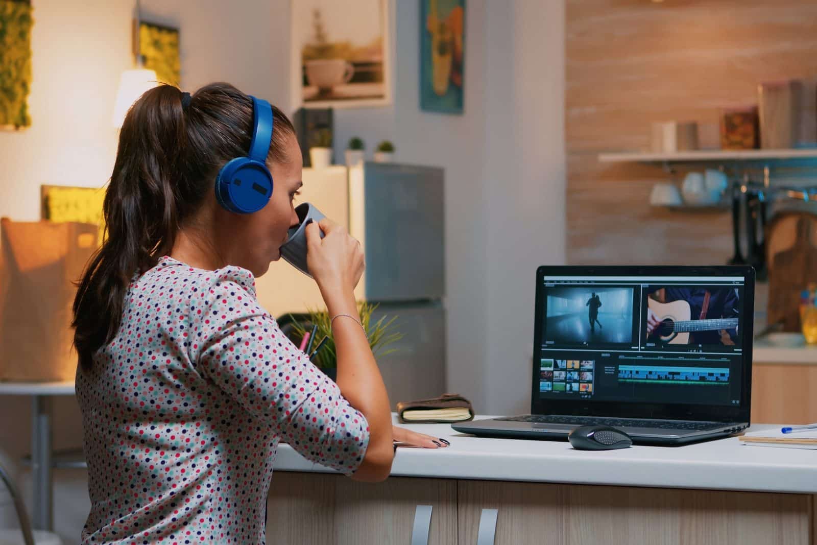 Woman video editor with headset working with footage and sound sitting in home kitchen. Woman videographer editing audio film montage on professional laptop sitting on desk in midnight.